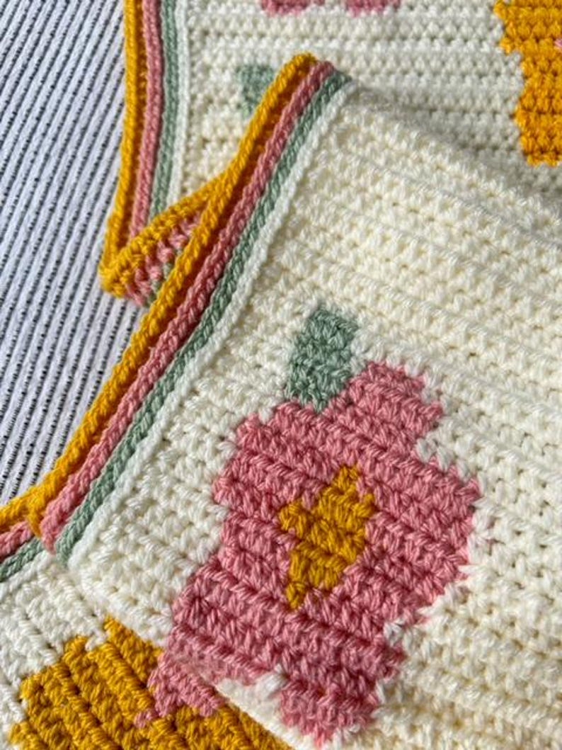 May include: A crocheted blanket with a floral pattern in shades of pink, yellow, and green. The blanket has a white background and a border of yellow, pink, and green.