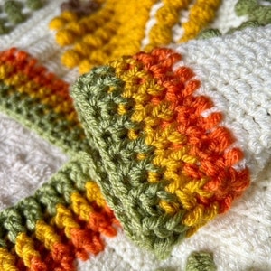 May include: Close-up of a crocheted blanket with a colorful striped border. The blanket is made with white, green, yellow, and orange yarn.