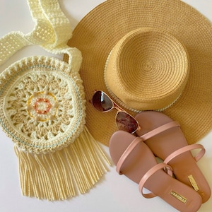 May include: A crocheted bag with a fringe, a straw hat, and a pair of brown sandals. The bag has a circular design with a beaded center.