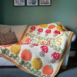 Puede incluir: Una manta de crochet con fondo blanco y detalles coloridos. La manta presenta un dise&ntilde;o de calabazas, setas y hojas. El texto "AUTUMN DAYS" est&aacute; cosido en la manta.