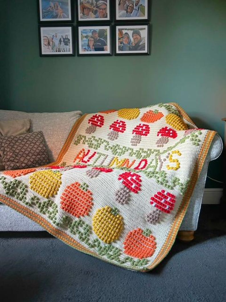 Puede incluir: Una manta de crochet con fondo blanco y detalles coloridos. La manta presenta un dise&ntilde;o de calabazas, setas y hojas. El texto "AUTUMN DAYS" est&aacute; cosido en la manta.