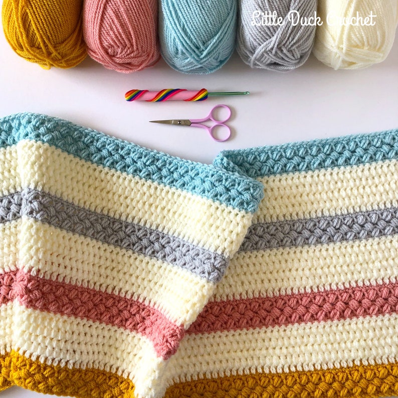 May include: A striped crochet blanket in yellow, pink, blue, grey and white. The blanket is folded in half and the yarn balls used to make the blanket are in the background.