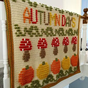 Puede incluir: Una manta de crochet con fondo crema y un dise&ntilde;o colorido que incluye las palabras "AUTUMN DAYS" en hilo rojo, naranja y amarillo. El dise&ntilde;o tambi&eacute;n incluye setas y calabazas en varios tonos de naranja, rojo y amarillo.