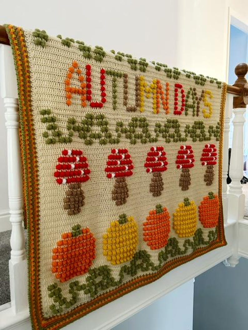 Puede incluir: Una manta de crochet con fondo crema y un dise&ntilde;o colorido que incluye las palabras "AUTUMN DAYS" en hilo rojo, naranja y amarillo. El dise&ntilde;o tambi&eacute;n incluye setas y calabazas en varios tonos de naranja, rojo y amarillo.