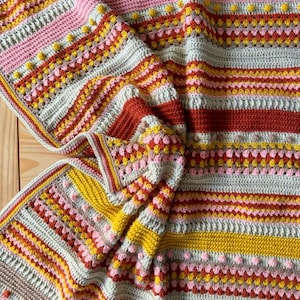 Puede incluir: Una manta de ganchillo hecha a mano con un diseño a rayas colorido. La manta presenta rayas rosas, amarillas, naranjas y grises. La manta está hecha con una variedad de puntadas, incluyendo bolitas y filas texturizadas. La manta está doblada y drapeada.