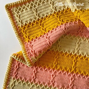 May include: A crocheted blanket with stripes of beige, yellow, and pink. The blanket is folded in half, showing the different colours and textures. The text "Little Duck Crochet" is visible in the top right corner of the image.