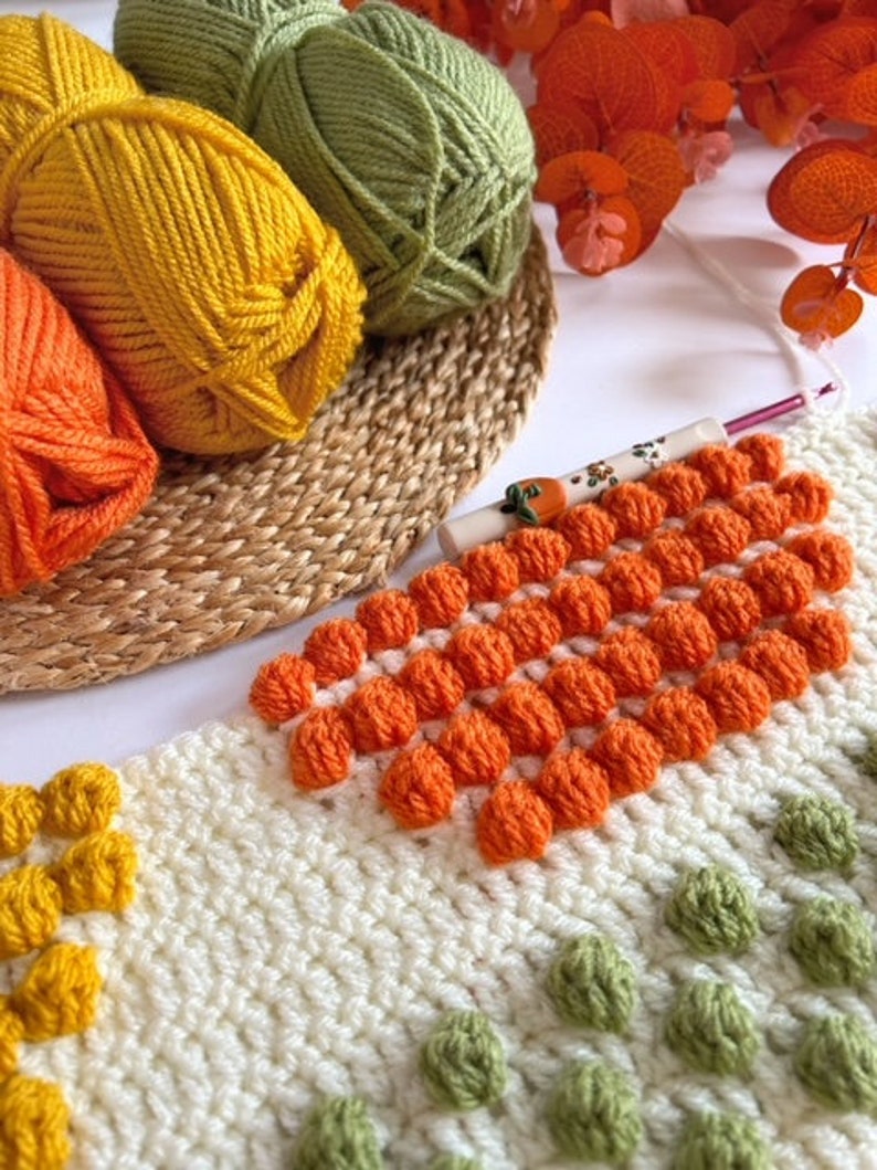 May include: A close-up of a crocheted blanket with a variety of colors and textures. The blanket features a pattern of orange, green, and yellow yarn. The blanket is being worked on with a crochet hook.