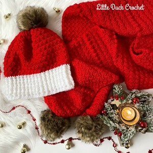 May include: Red and white crocheted hat with a brown faux fur pom pom, and a matching red crocheted scarf with a white trim. The hat and scarf are on a white fluffy surface with a small Christmas tree and a lit candle in the background.
