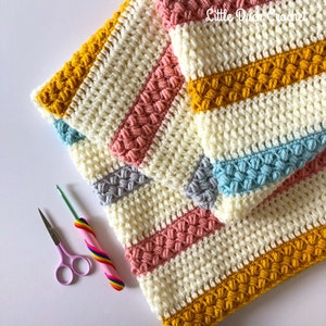 May include: A crocheted blanket with stripes of yellow, pink, blue, and grey. The blanket is made with a textured stitch that creates a bumpy surface. The blanket is folded in half and there are two crochet hooks and a pair of scissors on the white surface.