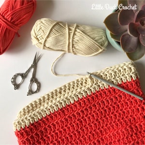 May include: A close-up of a crochet project in progress. The project is a two-toned, striped crochet bag with a cream colored top and a coral colored bottom. The bag is being worked on with a crochet hook and cream colored yarn. There are two pairs of small scissors and a ball of cream colored yarn in the background.