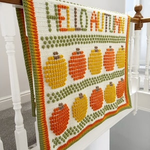 May include: A white crochet blanket with a green, orange, and yellow border. The blanket features the words "Hello Autumn" in brown yarn and a pattern of orange and yellow pumpkins.