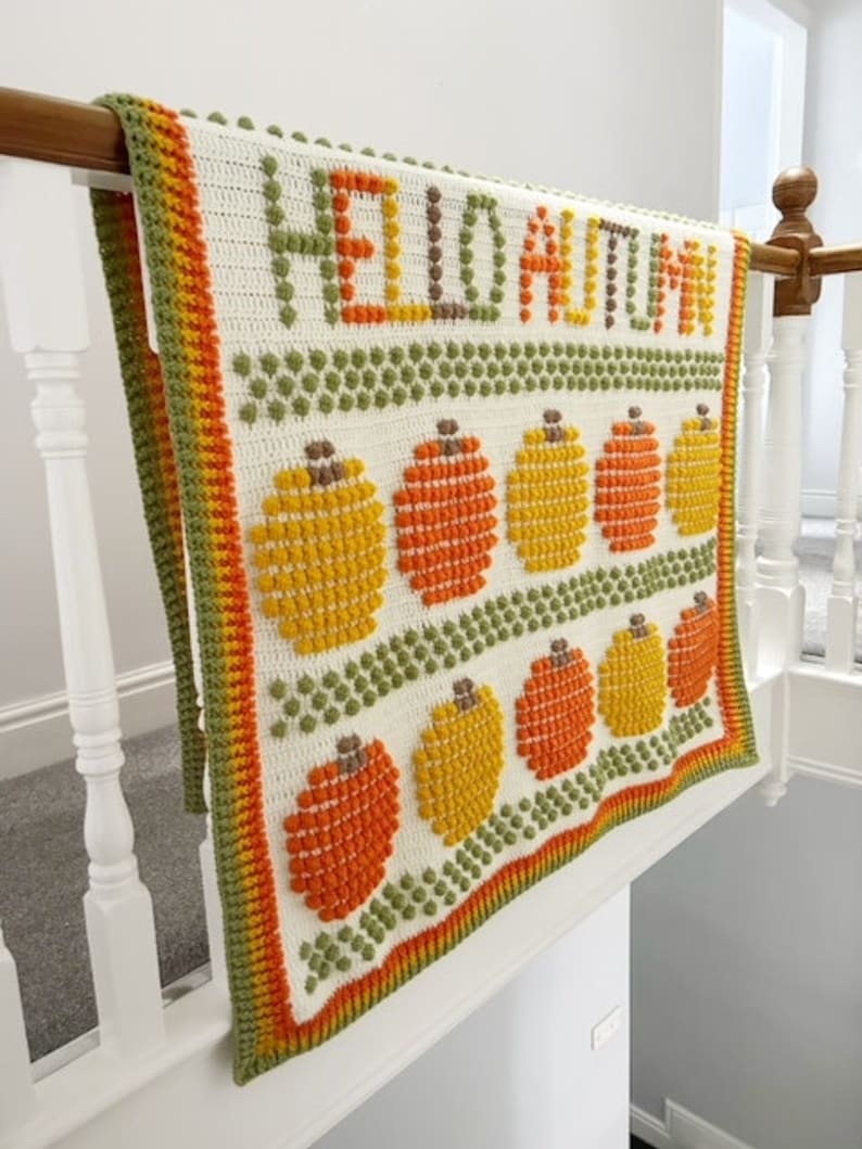 May include: A white crochet blanket with a green, orange, and yellow border. The blanket features the words "Hello Autumn" in brown yarn and a pattern of orange and yellow pumpkins.