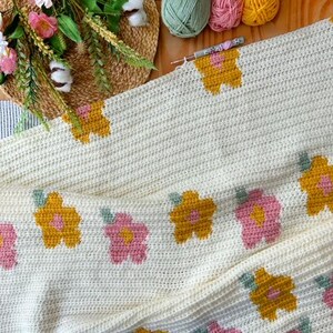 May include: A white crochet blanket with a repeating pattern of pink and yellow flowers. The blanket is folded in half and has a crocheted border.