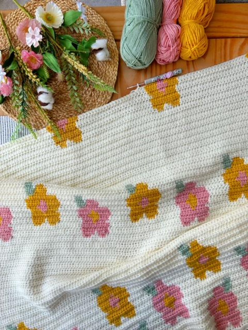 May include: A white crochet blanket with a repeating pattern of pink and yellow flowers. The blanket is folded in half and has a crocheted border.