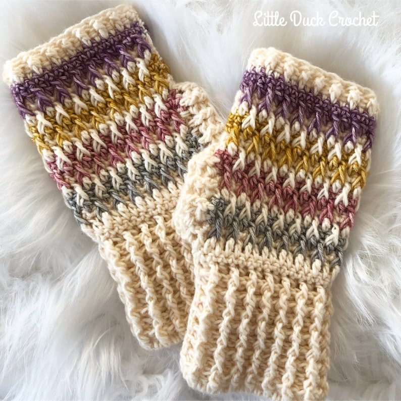 May include: A pair of crocheted fingerless gloves with a striped pattern in shades of purple, pink, gold, and gray. The gloves are made with a cream-colored yarn and have a textured stitch.