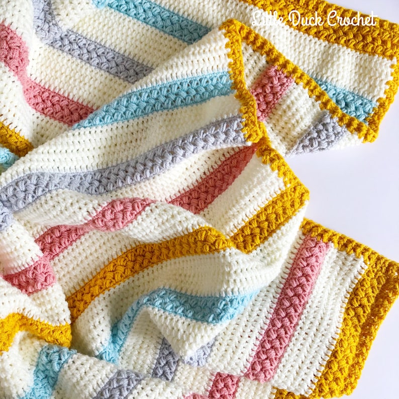 May include: A crocheted baby blanket with stripes of white, grey, pink, blue, and yellow. The blanket has a textured stitch and a yellow border.
