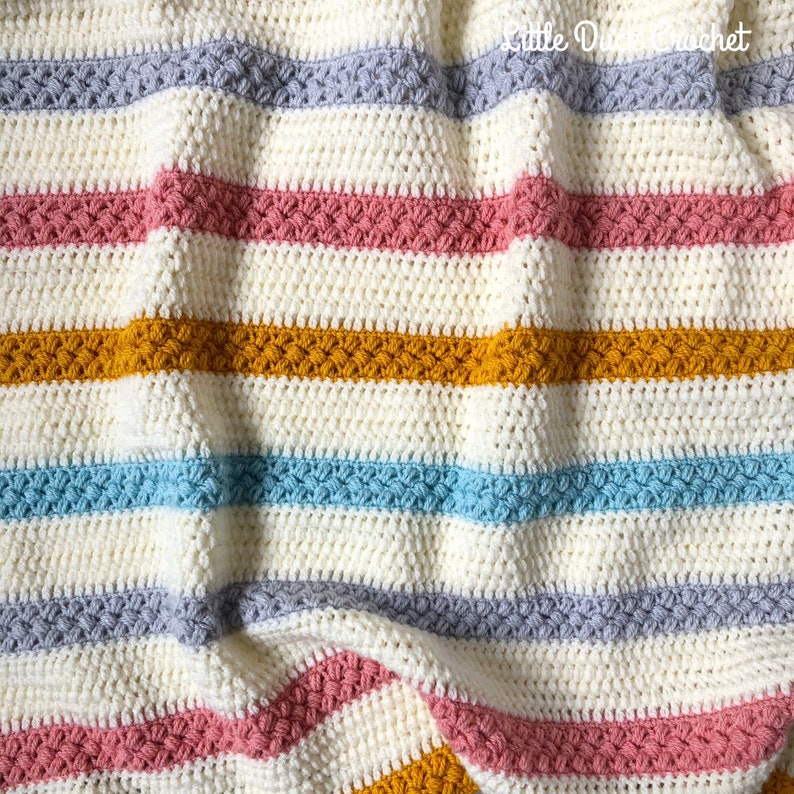 May include: A striped crochet blanket with a textured stitch pattern. The blanket is made in shades of white, grey, pink, blue, and yellow.