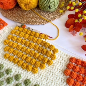 May include: A close-up of a crocheted blanket with squares of different colors. The squares are made with a bobble stitch and feature yellow, orange, and green yarn.