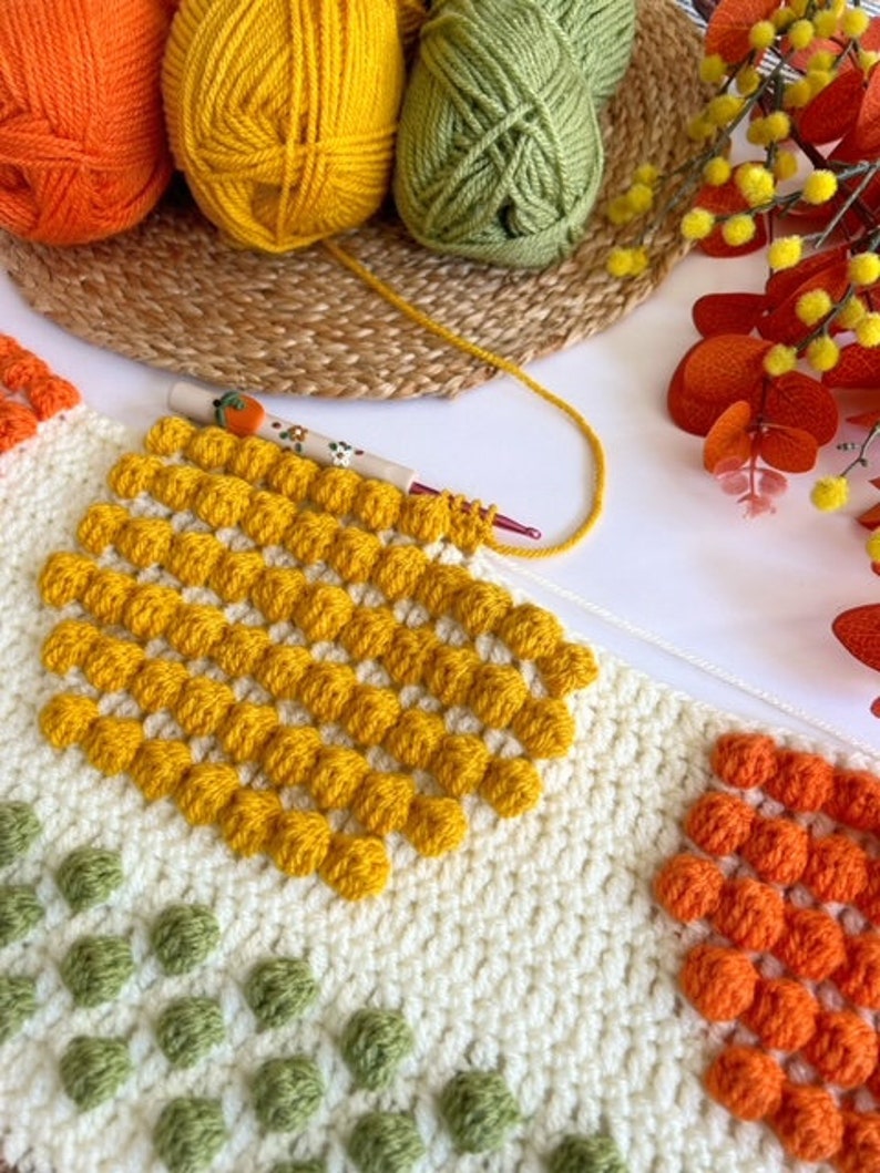 May include: A close-up of a crocheted blanket with squares of different colors. The squares are made with a bobble stitch and feature yellow, orange, and green yarn.