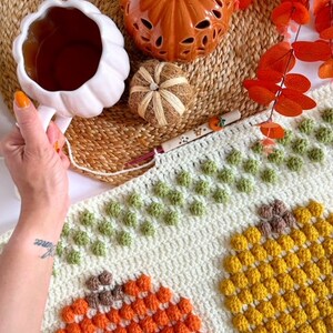 May include: A white crocheted blanket with a textured pumpkin design. The blanket features three different colored pumpkins: orange, yellow, and brown. The pumpkins are made with a bobble stitch.