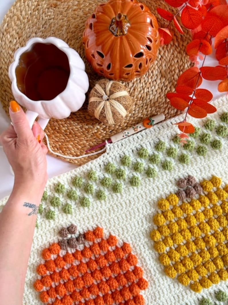May include: A white crocheted blanket with a textured pumpkin design. The blanket features three different colored pumpkins: orange, yellow, and brown. The pumpkins are made with a bobble stitch.
