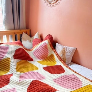 May include: A crocheted blanket with a geometric pattern of pink, yellow, and red triangles on a white background. The blanket is draped over a bed with pillows.