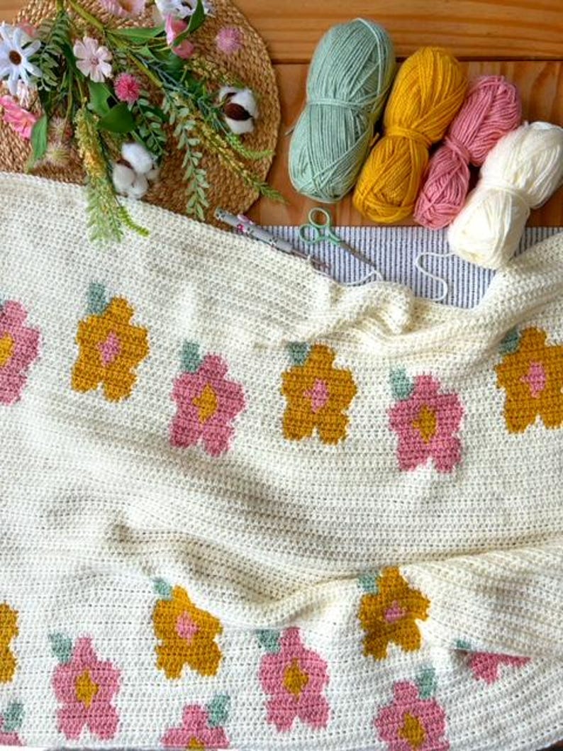 May include: A white crocheted blanket with a floral pattern. The blanket features pink, yellow, and green flowers. The blanket is folded in half and is lying on a wooden surface. There are balls of yarn in various colors in the background.