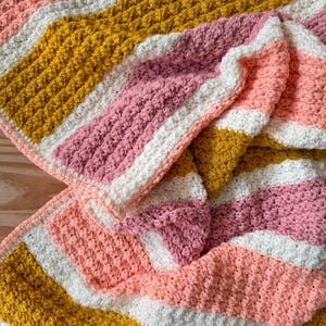 May include: A crocheted blanket featuring a repeating striped pattern in shades of mustard yellow, cream white, soft pink, and coral orange. The textile displays a textured, bumpy stitch pattern throughout, creating a consistent, tactile surface.