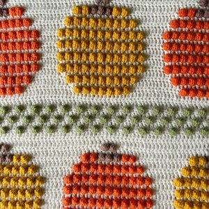 May include: A crocheted blanket with a pattern of orange and yellow pumpkins. The pumpkins are made with a bobble stitch and are surrounded by a white background.