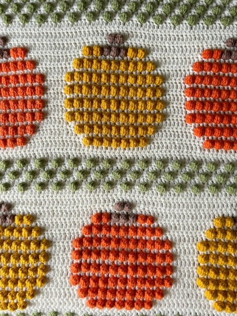 May include: A crocheted blanket with a pattern of orange and yellow pumpkins. The pumpkins are made with a bobble stitch and are surrounded by a white background.