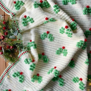 May include: A cream-coloured, crocheted blanket with a repeating pattern of green holly leaves and red berry accents. The blanket has a textured appearance and a decorative border with red and green stripes, ideal for festive home decor.