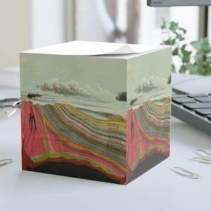 May include: A cube-shaped notepad featuring a vintage geological illustration. The artwork depicts layered rock formations in shades of red, brown, yellow, and green, under a sky with clouds. The notepad sits on a white surface with paper clips.