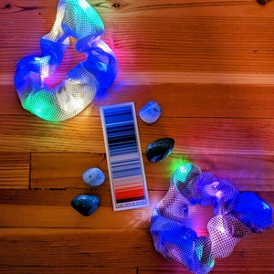 Pu&ograve; includere: Due scrunchies per capelli colorati con un design a rete, illuminati da luci LED. Gli scrunchies sono su una superficie di legno con una cartella colori e pietre blu e verdi.