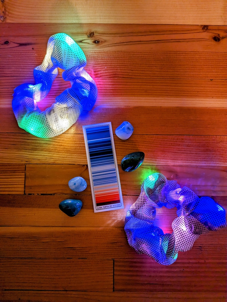 Pu&ograve; includere: Due scrunchies per capelli colorati con un design a rete, illuminati da luci LED. Gli scrunchies sono su una superficie di legno con una cartella colori e pietre blu e verdi.