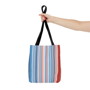 May include: A tote bag with a colorful vertical stripe pattern in shades of blue, orange, and red. The bag has black handles and is held up by a hand with red nail polish. The bag is made of a sturdy material.