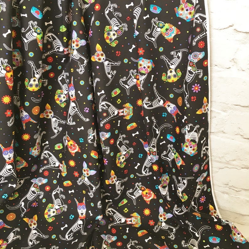 May include: Black fabric with a repeating pattern of white skeleton dogs with colorful flowers and bones. The dogs are in various poses, some are standing, some are sitting, and some are running.