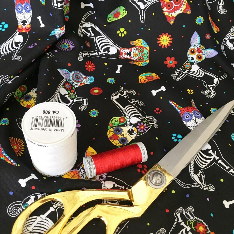 May include: Black fabric with a repeating pattern of colorful sugar skull dogs. The fabric is surrounded by white thread, red thread, and gold scissors. The thread spools have labels that read "Made in Germany" and "100% Viscose".