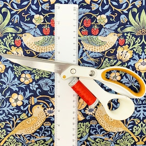 May include: White and gold-handled scissors, a spool of red thread, and a ruler rest on a patterned fabric. The fabric features a dark blue background with a repeating design of birds, strawberries, and floral elements.