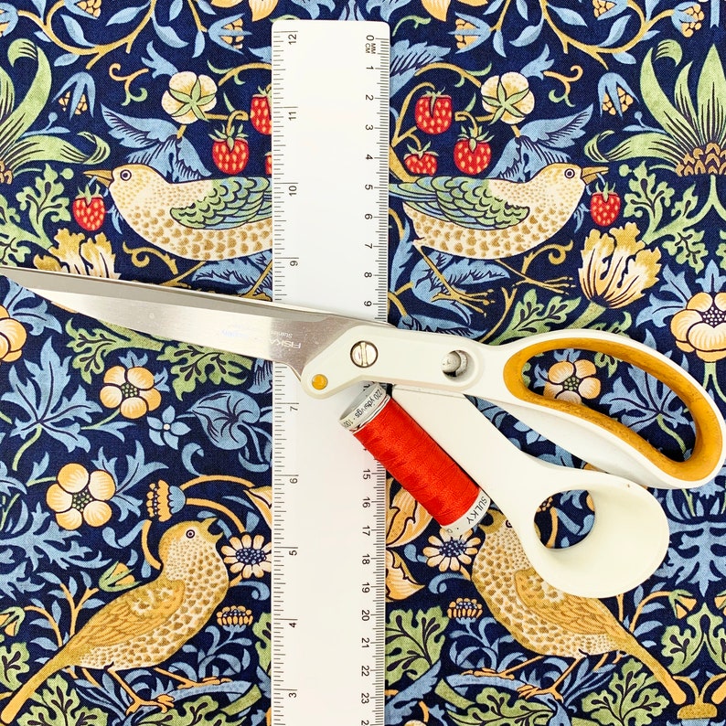 May include: White and gold-handled scissors, a spool of red thread, and a ruler rest on a patterned fabric. The fabric features a dark blue background with a repeating design of birds, strawberries, and floral elements.
