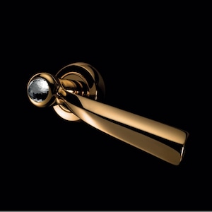 Könnte beinhalten: Ein goldfarbener Türgriff mit Kristallakzent. Der Griff hat ein geschwungenes Design und eine polierte Oberfläche, vor schwarzem Hintergrund. Der Kristall ist facettiert und reflektiert Licht.
