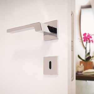 Könnte beinhalten: Ein moderner, verchromter Türgriff und eine quadratische Schlüssellochabdeckung sind an einer weißen Tür angebracht. Der Griff hat ein schlankes, rechteckiges Design. Die Tür öffnet sich zu einem Badezimmer mit Waschbecken und rosa Blumenarrangement.