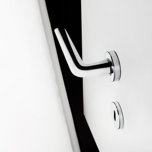 Puede incluir: Una manija de puerta cromada con un diseño de palanca y una tapa redonda y plana para la cerradura. La manija está montada en una puerta blanca.