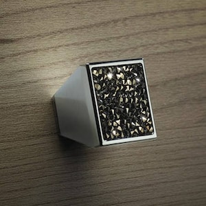 Könnte beinhalten: Ein quadratischer Schrank-Knopf mit einer silbernen Basis und einer schwarzen kristallartigen Oberfläche.
