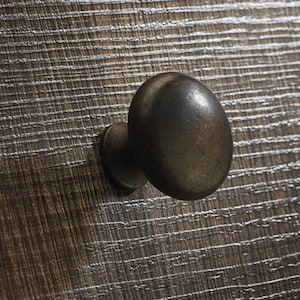 Könnte beinhalten: Nahaufnahme einer dunkelbraunen Holzoberfläche mit einem strukturierten, linearen Muster. Ein kleiner, abgerundeter, bronzefarbener Knopf ist am Holz befestigt. Der Knopf ist oval geformt und hat einen passenden Bronzesockel.