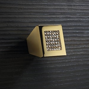 Könnte beinhalten: Ein goldfarbener Schrankgriff mit einer rechteckigen Fläche, in die kleine, klare Kristalle eingesetzt sind. Der Griff ist auf einer dunklen Holzmaserung montiert, was einen Kontrast erzeugt.