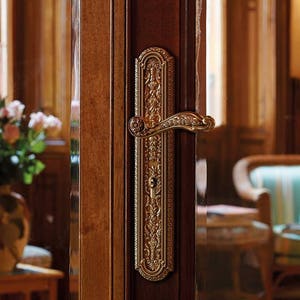 Könnte beinhalten: Ein goldener Türgriff mit aufwendigen floralen Mustern und einem gebogenen Hebel. Der Griff ist an einer Holztür montiert.