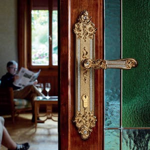May include: A gold door handle with a decorative design and a keyhole. The handle is made of metal and has a polished finish. The handle is mounted on a wooden door with a glass panel. The text "PATENTED DESIGN" is visible in the upper right corner of the image.