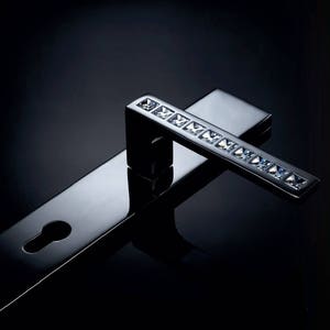 Puede incluir: Una manilla de puerta plateada con una palanca larga y rectangular adornada con una fila de adornos de cristal transparente. La manilla está diseñada para una sola puerta y tiene una cerradura.