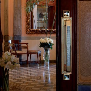 Peut inclure: Une poignée de porte dorée avec un design en cristal. La poignée est montée sur une porte en bois. La porte est ouverte et on peut voir une pièce avec un miroir, un vase de fleurs blanches et deux chaises.