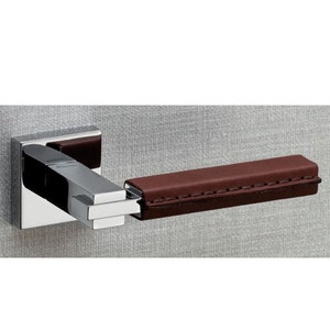 May include: A chrome door handle with a brown leather grip. The handle has a square base and a rectangular grip with stitching.